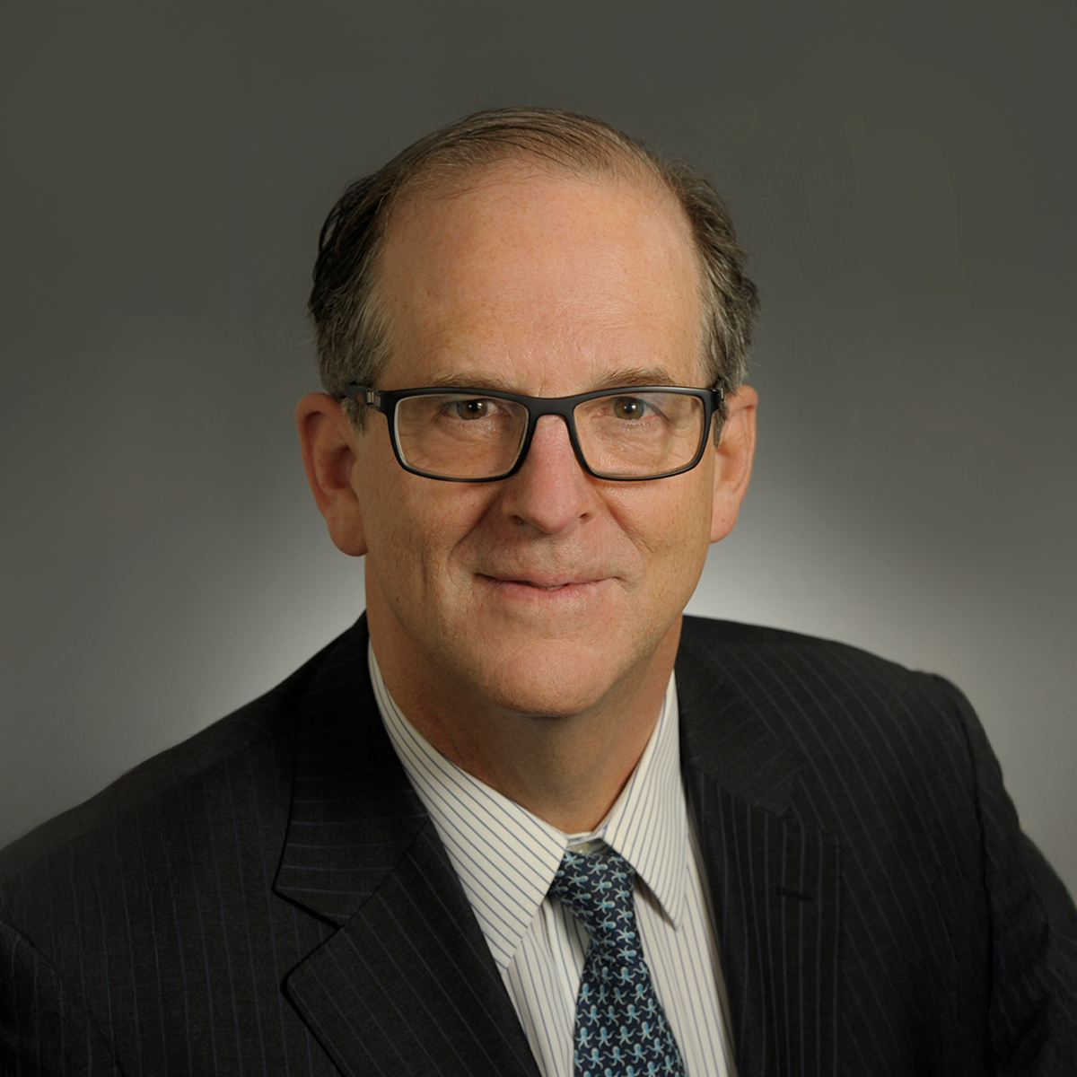 Portrait of a businessman wearing glasses and a tie, facing forward with a neutral expression against a plain background.