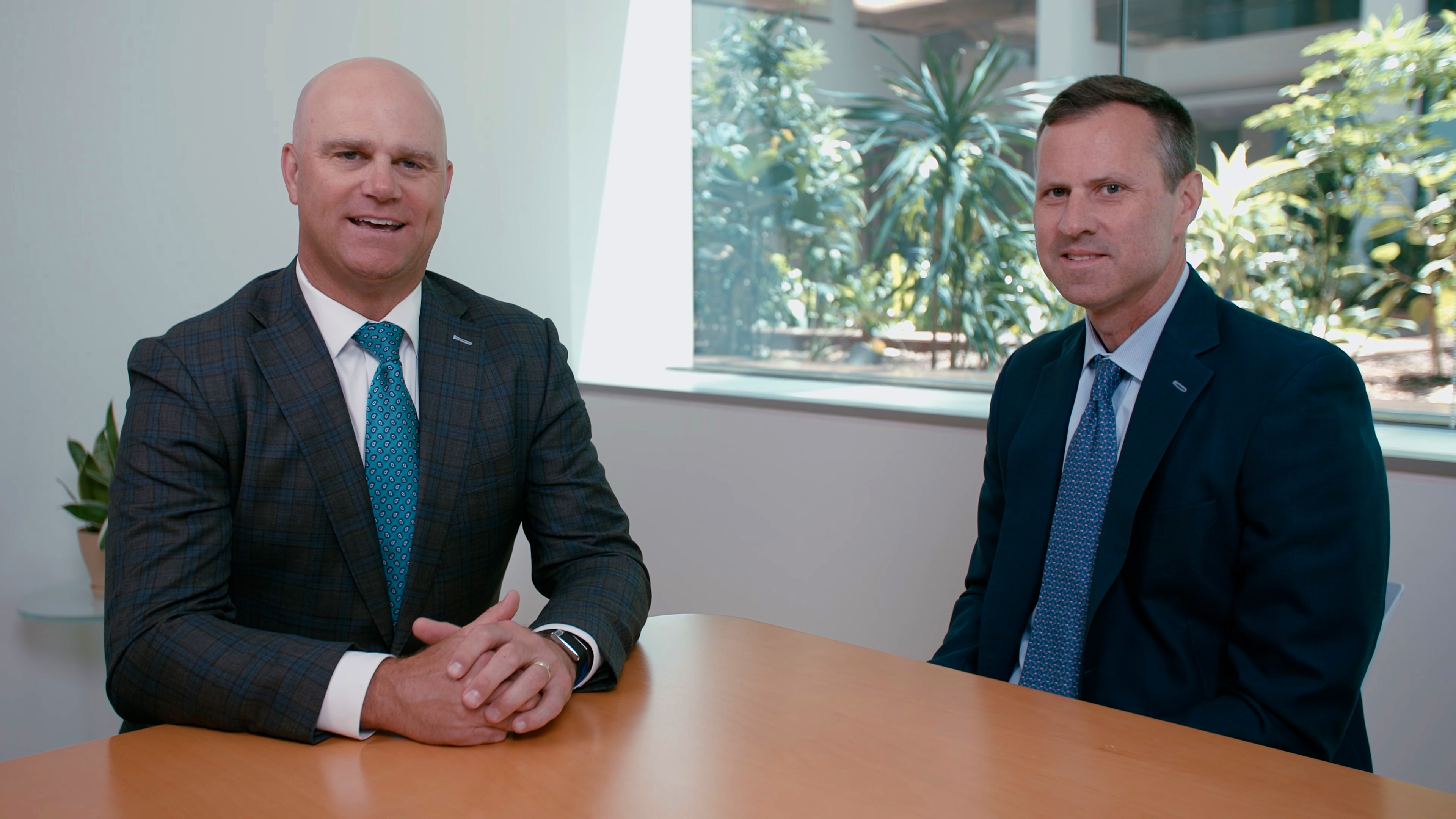 A smiling businessman stands confidently in a corporate office environment, surrounded by professional colleagues.