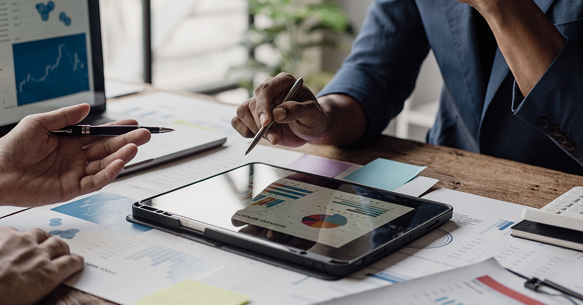Two people review financial charts on a tablet at a desk, holding pens over printed documents and notes.