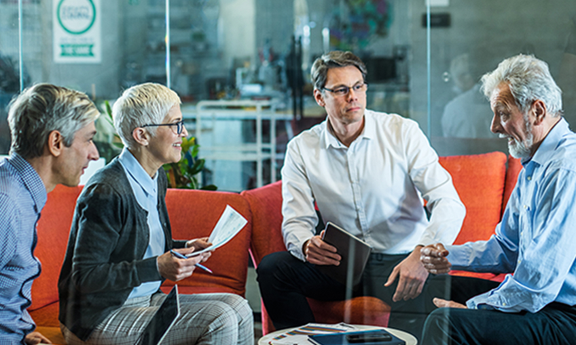 Four adults seated in a lounge area reviewing documents and discussing financial planning, with one person holding a tablet and another holding printed materials.