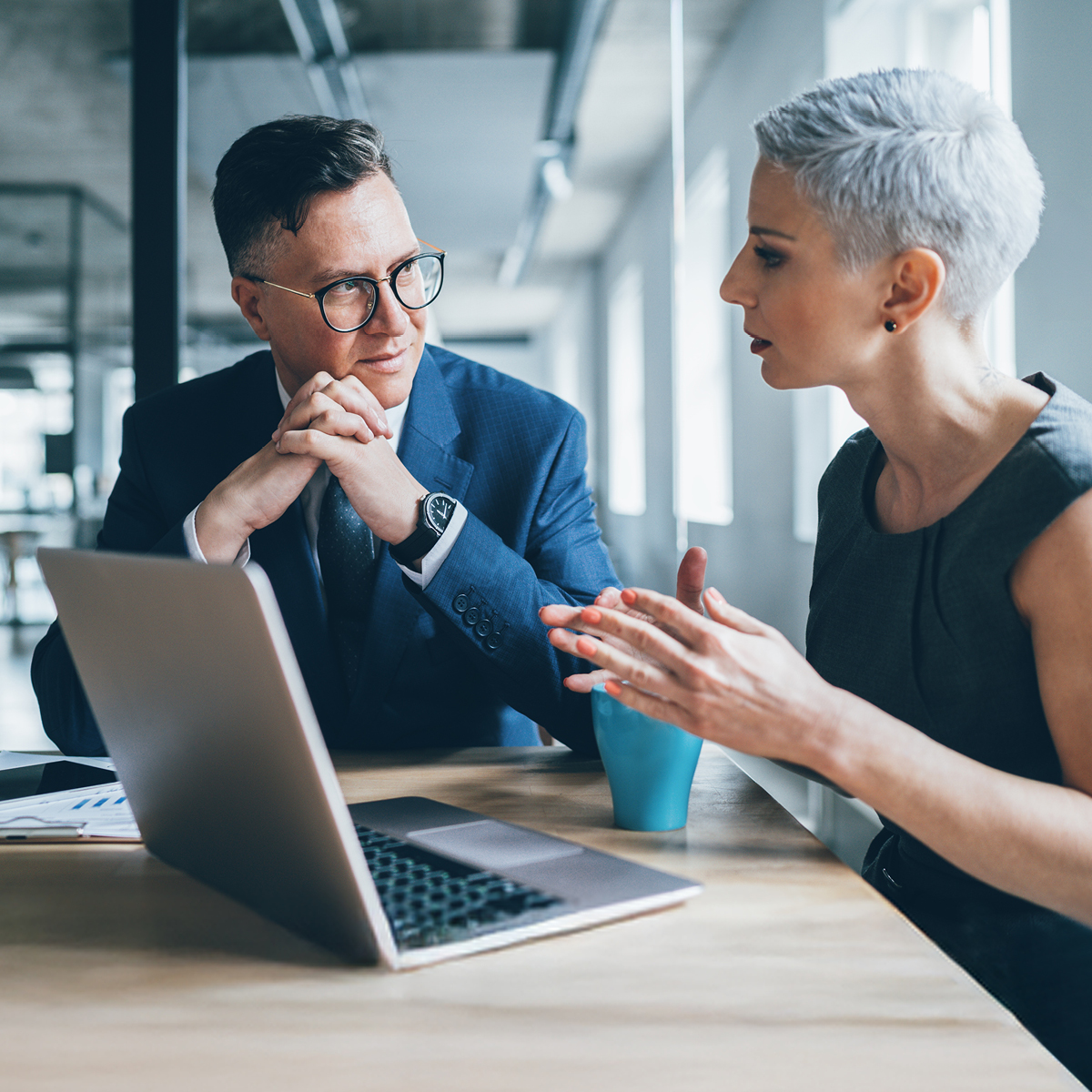 A woman and a man work collaboratively at a desk in a modern office, using a laptop and notebook for business tasks.