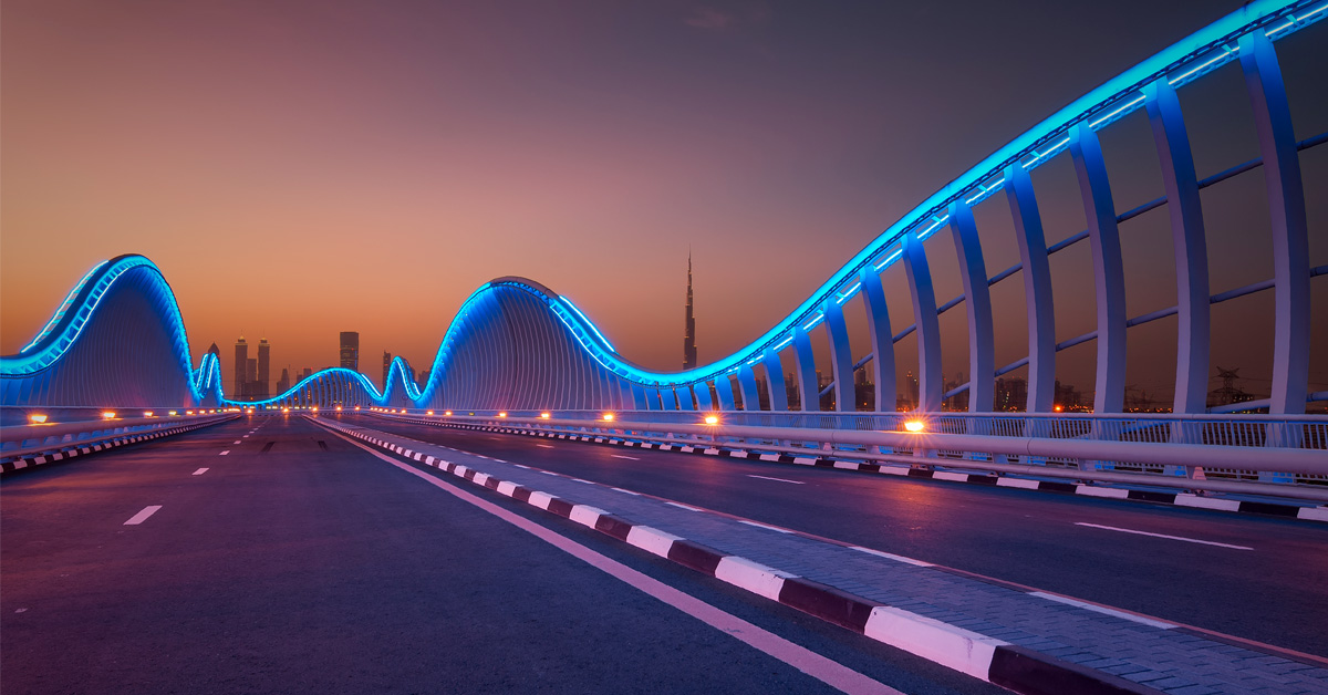 A vibrant nighttime cityscape featuring illuminated architecture, a lit bridge, and flowing highway traffic under a blue sky.